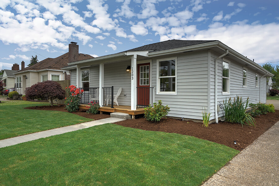 Photo of a grey one-story bungalow with grass and flowers in the front yard.